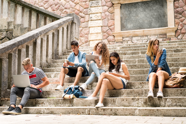 Group of attractive teenage students sitting on stone steps in front of university holding tablet, laptop and smart phones, reading or watching something.