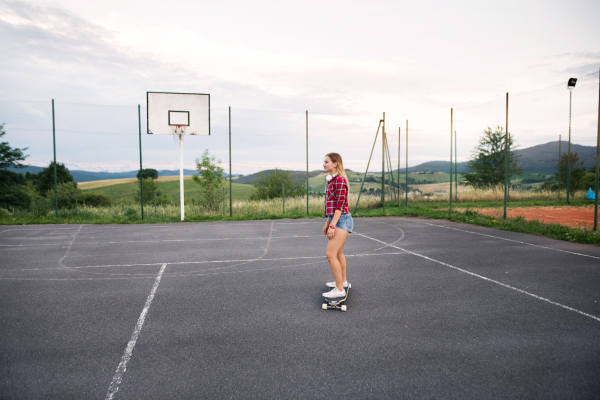 Attractive teenage student girl standing on skateboard outdoors on basketball playground.