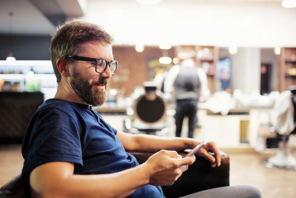 Handsome hipster man client visiting haidresser and hairstylist in barber shop, using smartphone when waiting.