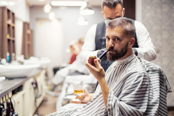 Handsome hipster man client visiting haidresser and hairstylist in barber shop, smoking a pipe.