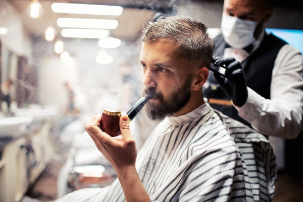 Handsome hipster man client visiting haidresser and hairstylist in barber shop, smoking a pipe.