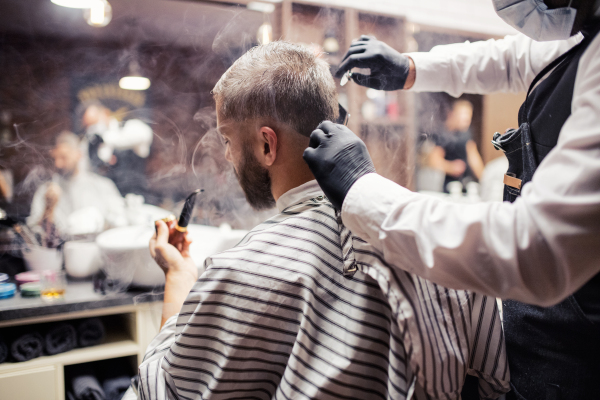 Handsome hipster man client visiting haidresser and hairstylist in barber shop, smoking a pipe.