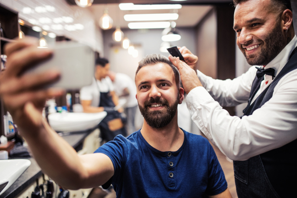 Handsome hipster man client visiting haidresser and hairstylist in barber shop, taking selfie with smartphone.