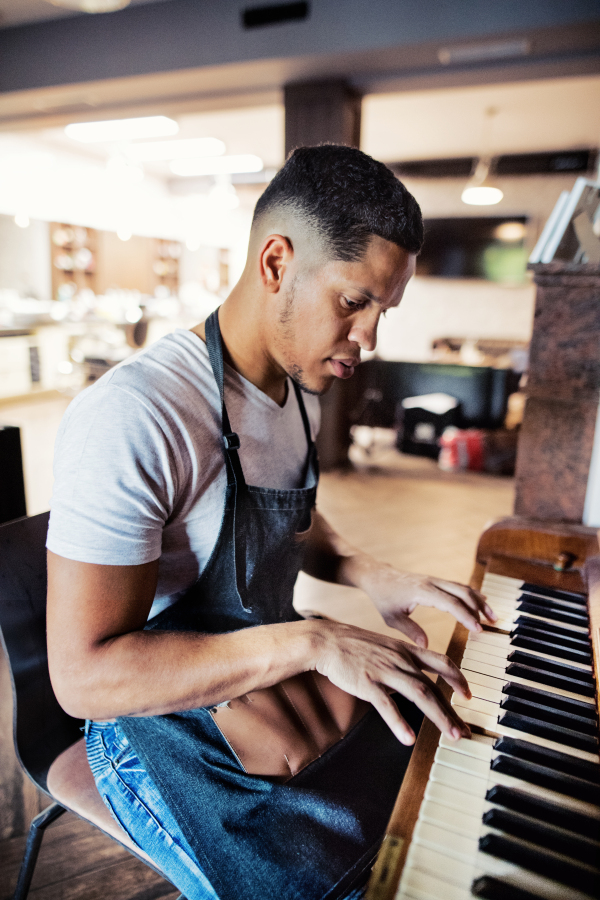 Young male hispanic haidresser and hairstylist sitting in barber shop, playing piano.