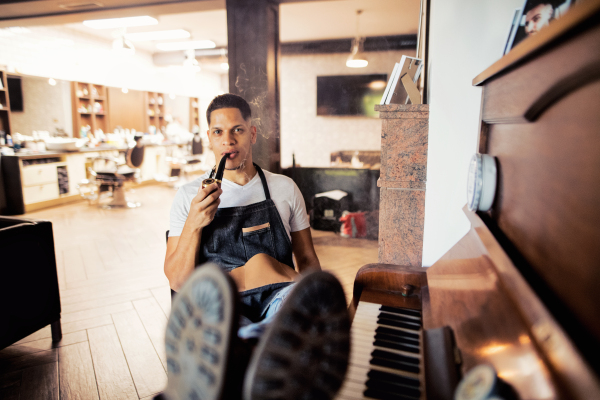 Young male hispanic haidresser and hairstylist sitting in barber shop, smoking a pipe and holding a drink.