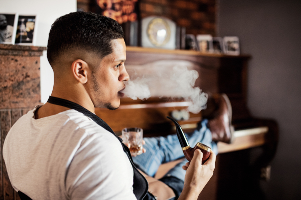 Young male hispanic haidresser and hairstylist sitting in barber shop, smoking a pipe and holding a drink.