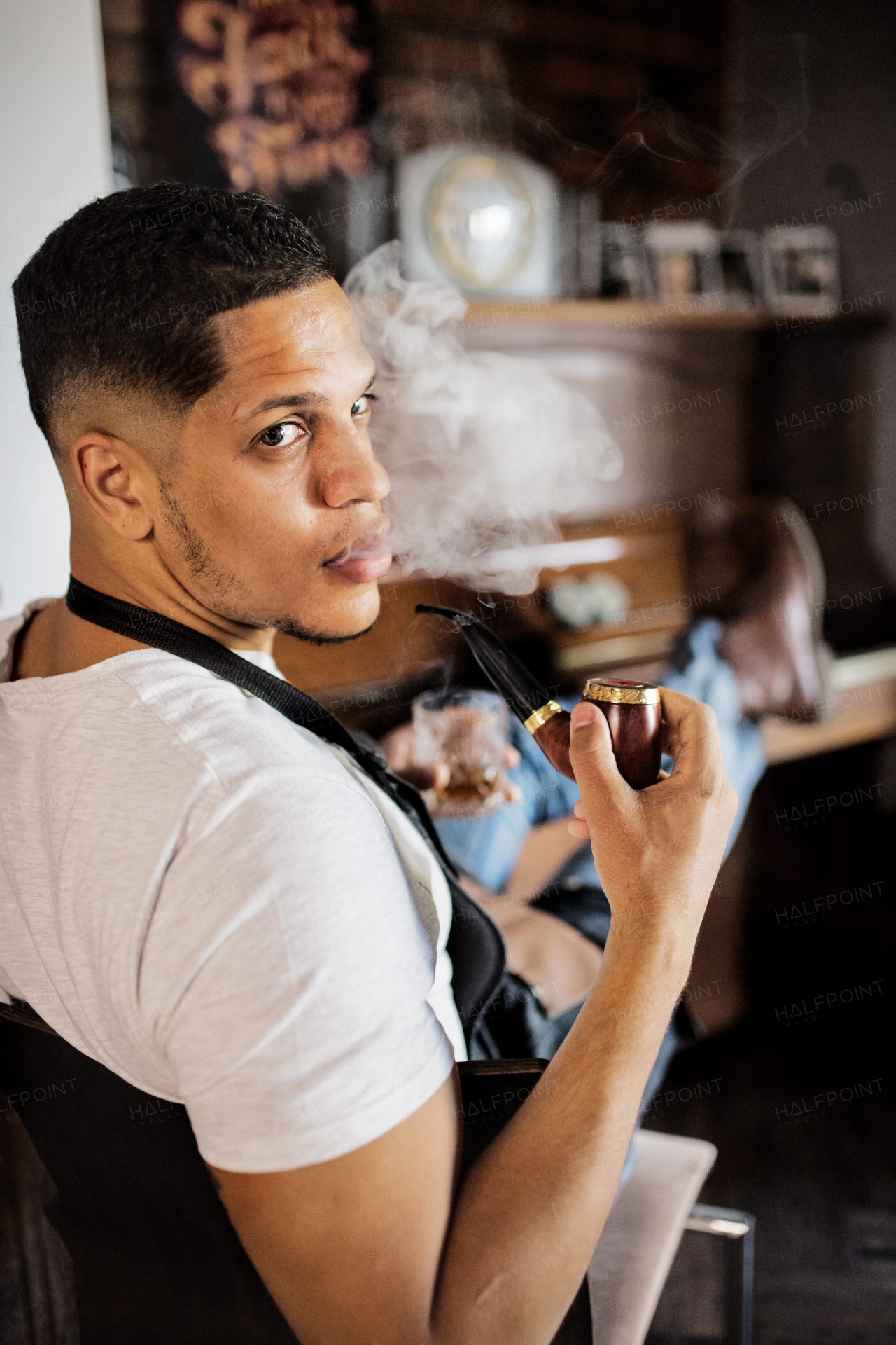 Young male hispanic haidresser and hairstylist sitting in barber shop, smoking a pipe and holding a drink.