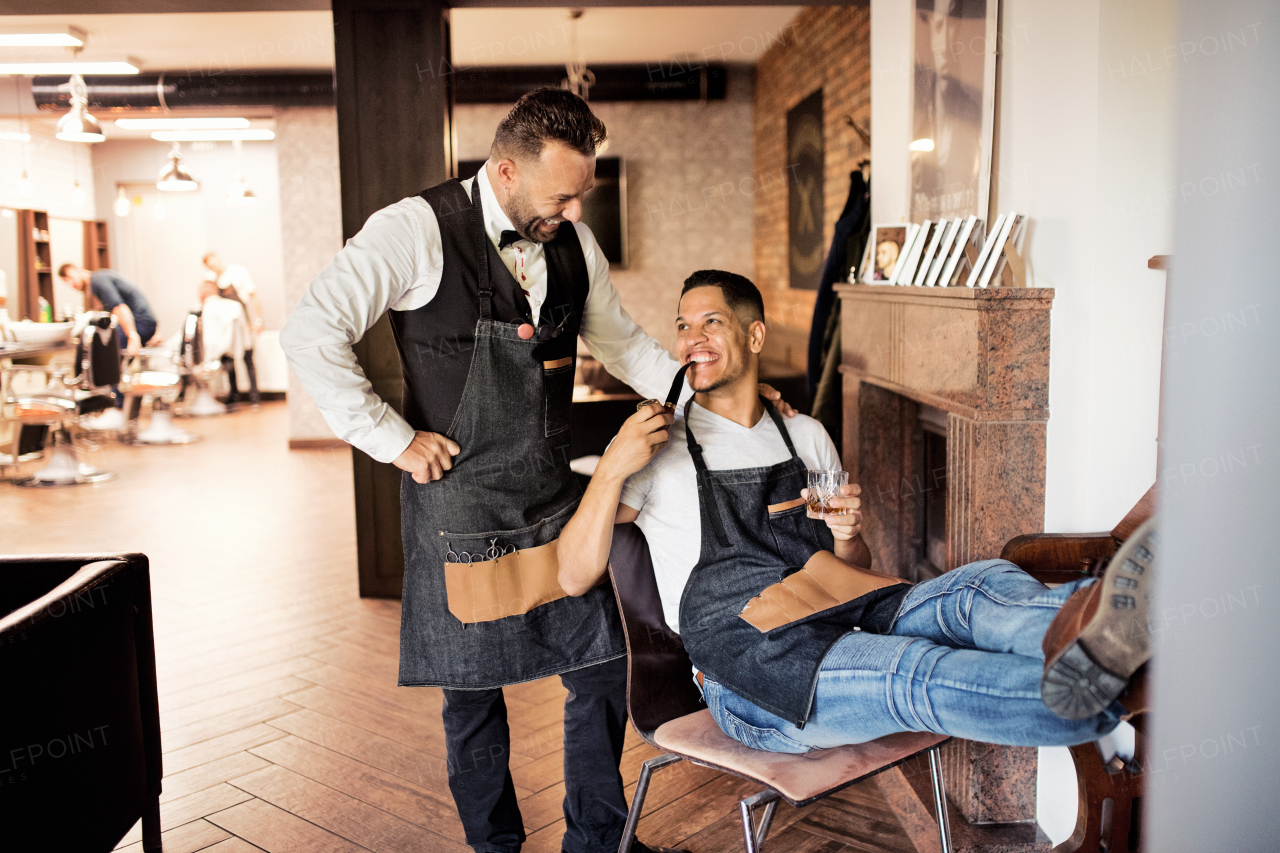 Two cheerful haidresser and hairstylist in barber shop, smoking a pipe and talking.