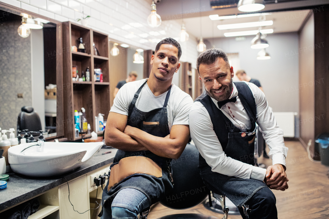 Two male haidressers and hairstylists sitting in barber shop, posing for a photograph.