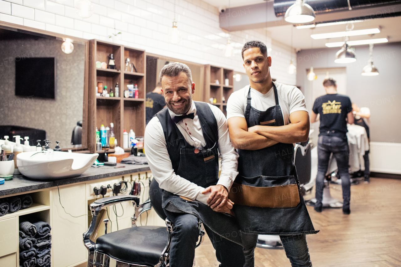 Two male haidressers and hairstylists sitting in barber shop, posing for a photograph.