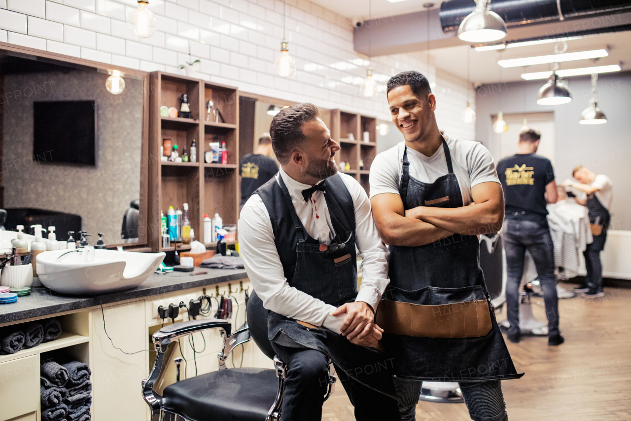 Two male haidressers and hairstylists sitting in barber shop, posing for a photograph.