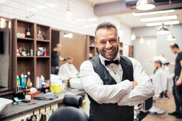 Young handsome haidresser and hairstylist standing in barber shop, arms crossed.