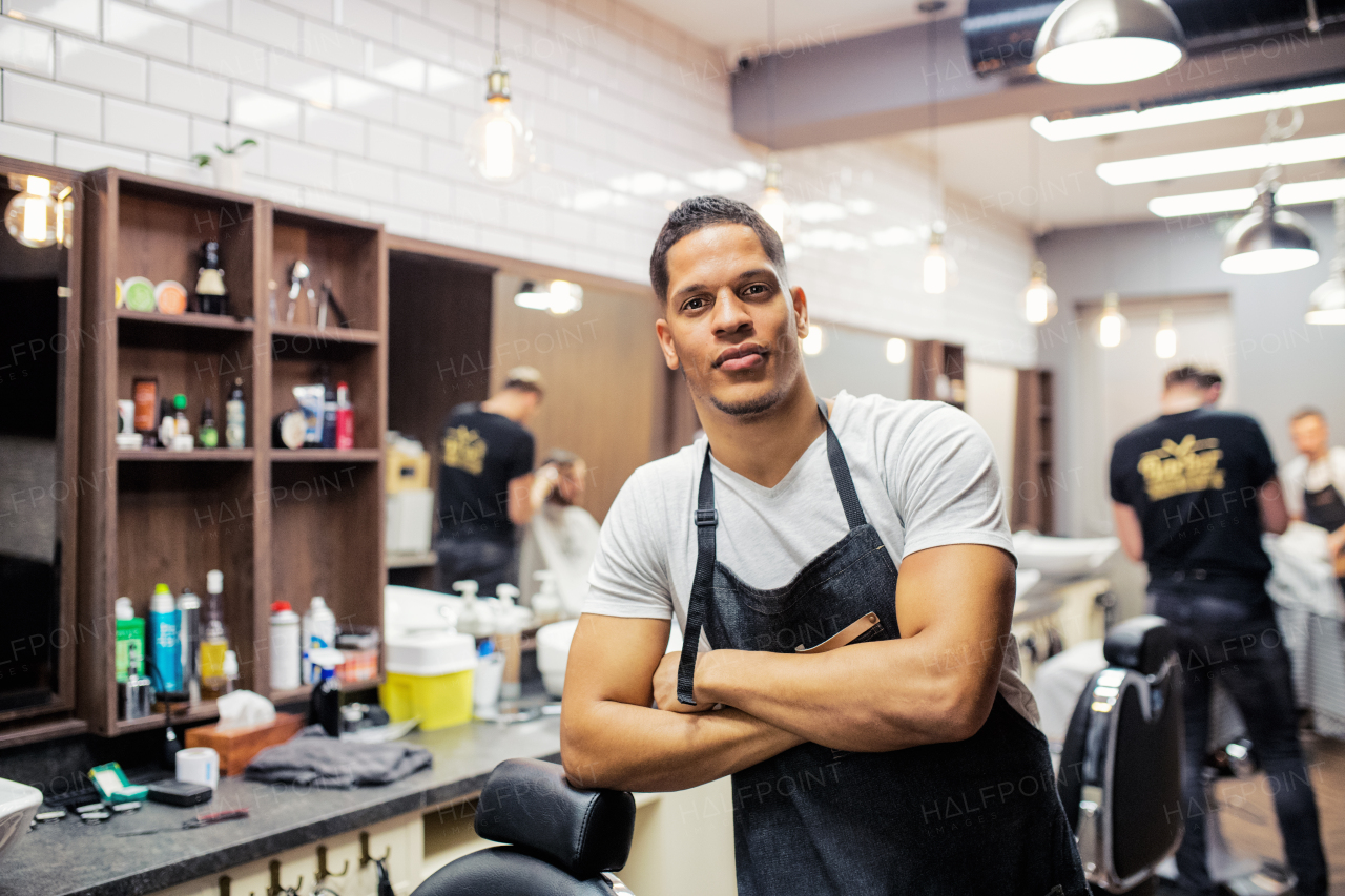 Young handsome hispanic haidresser and hairstylist standing in barber shop, arms crossed.