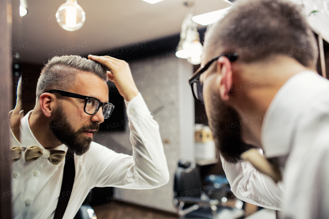 Handsome hipster man client with glasses looking in the mirror in barber shop.