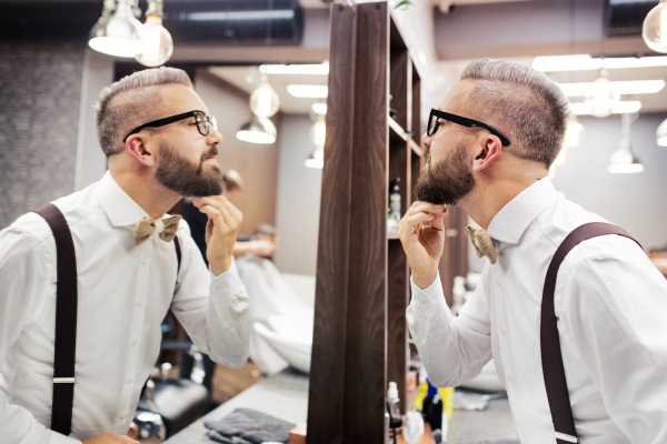 Handsome hipster man client with glasses looking in the mirror in barber shop.