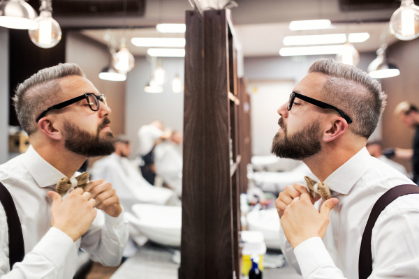 Handsome hipster man client with glasses looking in the mirror in barber shop.