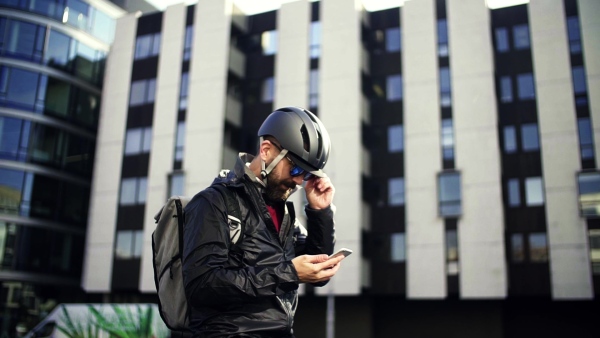 A portrait of male bicycle courier with backpack delivering packages in city, using smartphone. Slow motion.