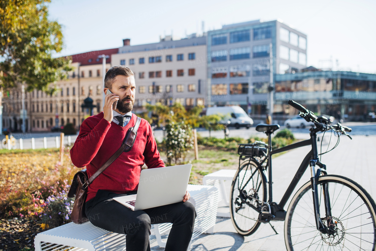 Hipster businessman commuter with electric bicycle sitting on bench, using laptop and smartphone.