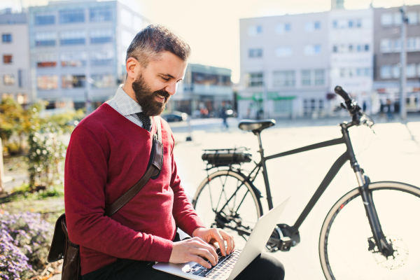 Hipster businessman commuter with bicycle on the way to work in city, sitting on a bench and using laptop.