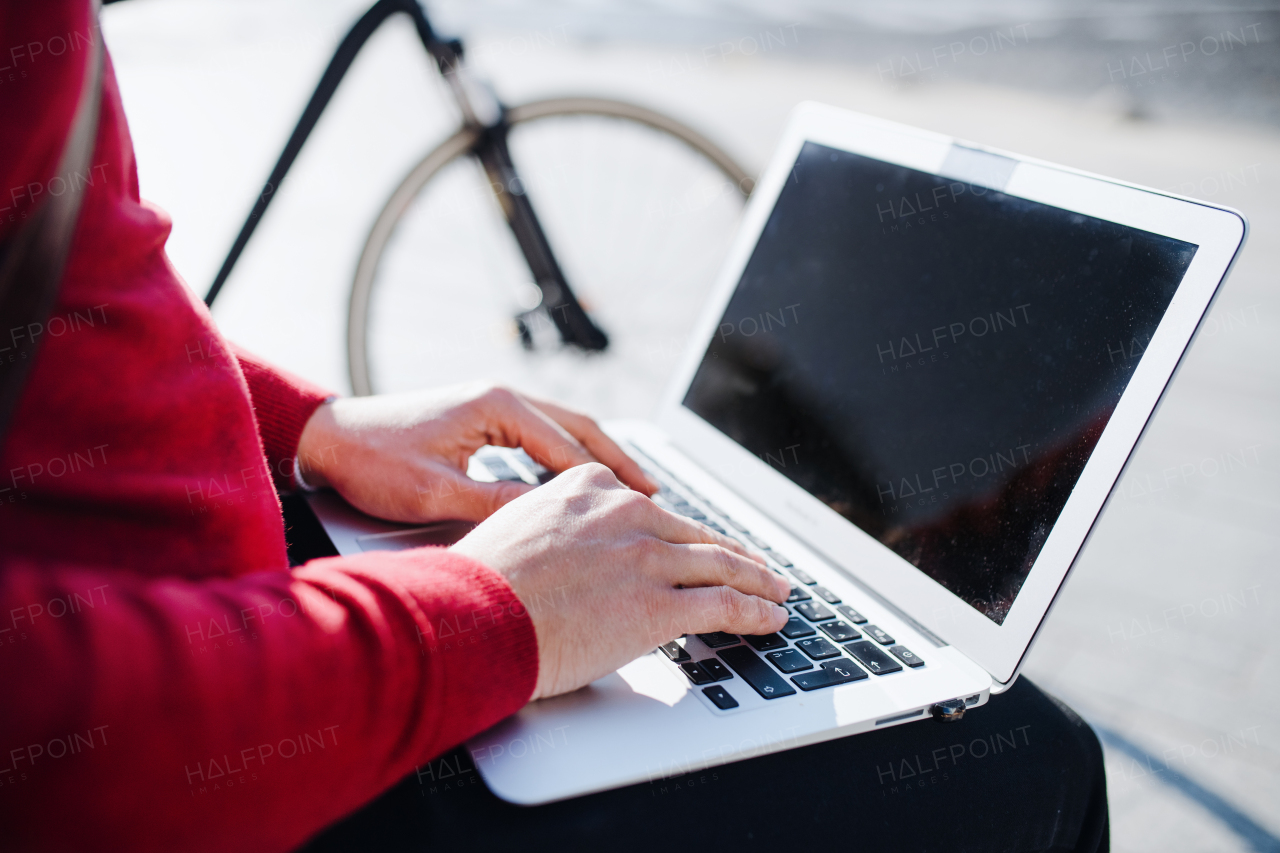 A midsection of businessman commuter with bicycle sitting on bench in city, using laptop. Copy space.