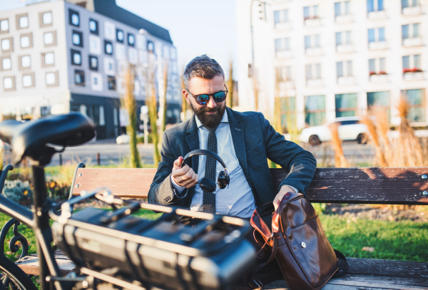 Businessman commuter with headphones and bicycle sitting on bench in city, resting.