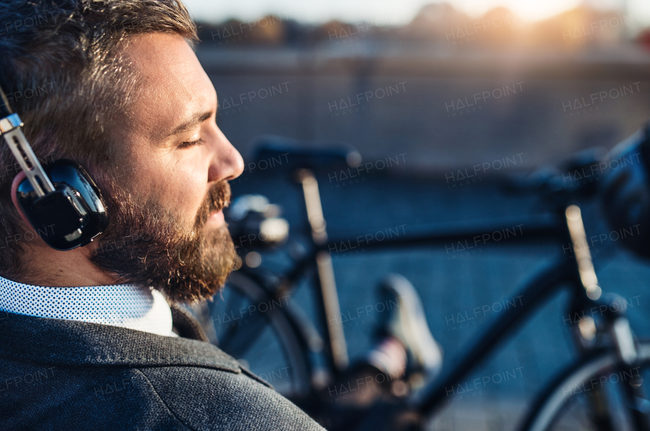 A close-up of businessman commuter with headphones and bicycle sitting on bench in city, listening to music. Copy space.