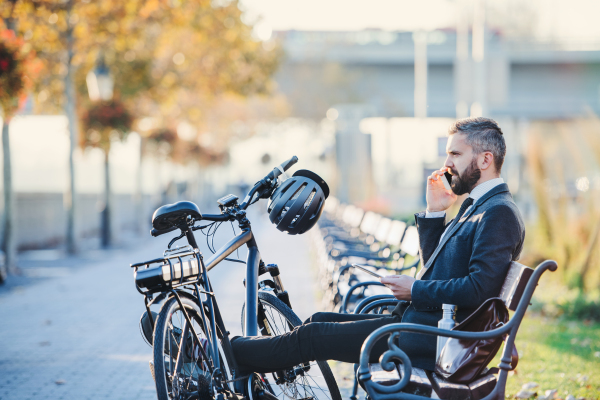 A side view of businessman commuter with smartphone and bicycle sitting on bench in city, making a phone call.