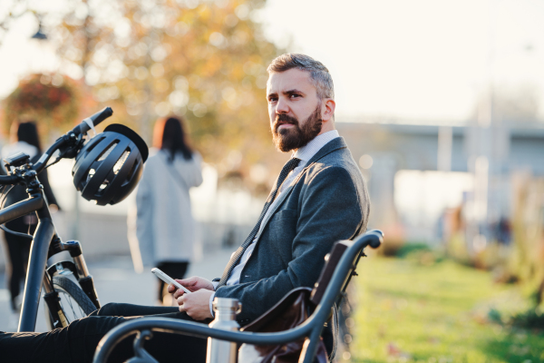 A side view of businessman commuter with smartphone and bicycle sitting on bench in city.