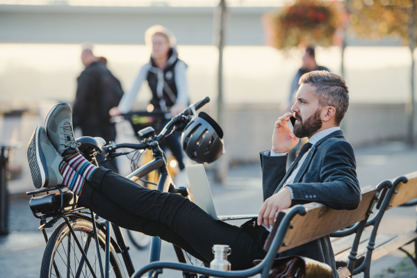 A side view of businessman commuter with smartphone and bicycle sitting on bench in city, making a phone call.