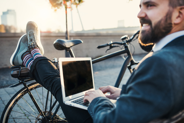 A businessman commuter with bicycle sitting on bench in city and using laptop when traveling home from work. Copy space.