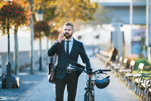 A businessman commuter with bicycle walking home from work in city, using smartphone.