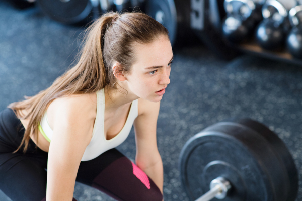 Beautiful young fit woman in gym lifting heavy barbell, flexing muscles