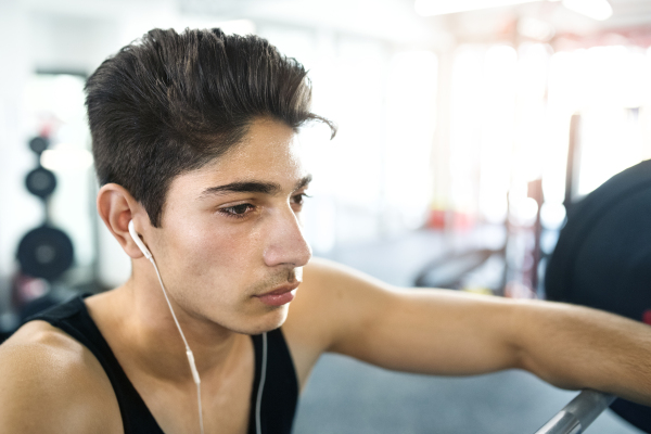 Hispanic fitness man in gym at the heavy barbell, resting, earphones in his ears, listening music