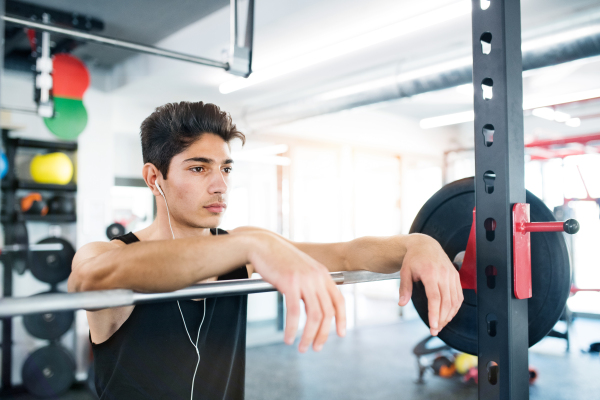 Hispanic fitness man in gym at the heavy barbell, resting, earphones in his ears, listening music