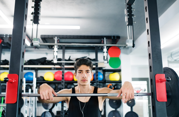 Hispanic fitness man in gym at the heavy barbell, resting, earphones in his ears, listening music