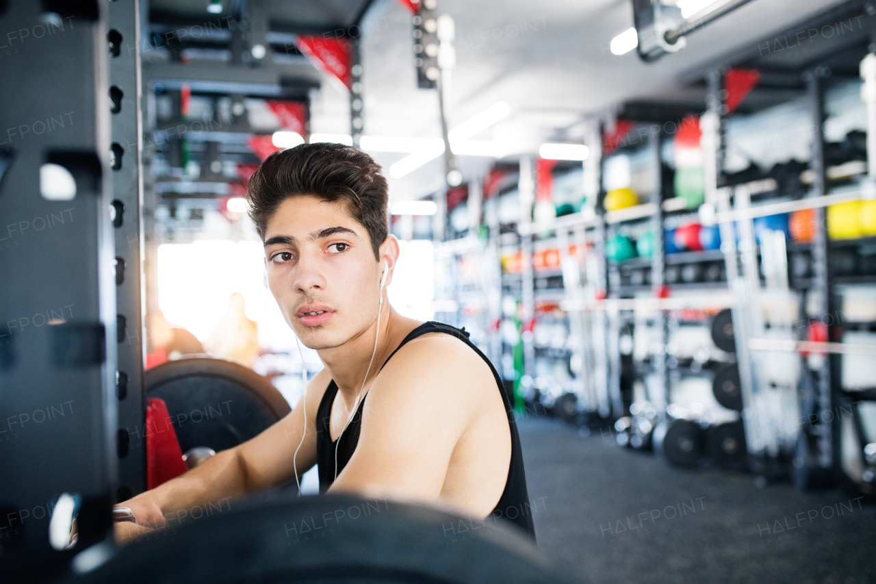 Hispanic fitness man in gym at the heavy barbell, resting, earphones in his ears, listening music