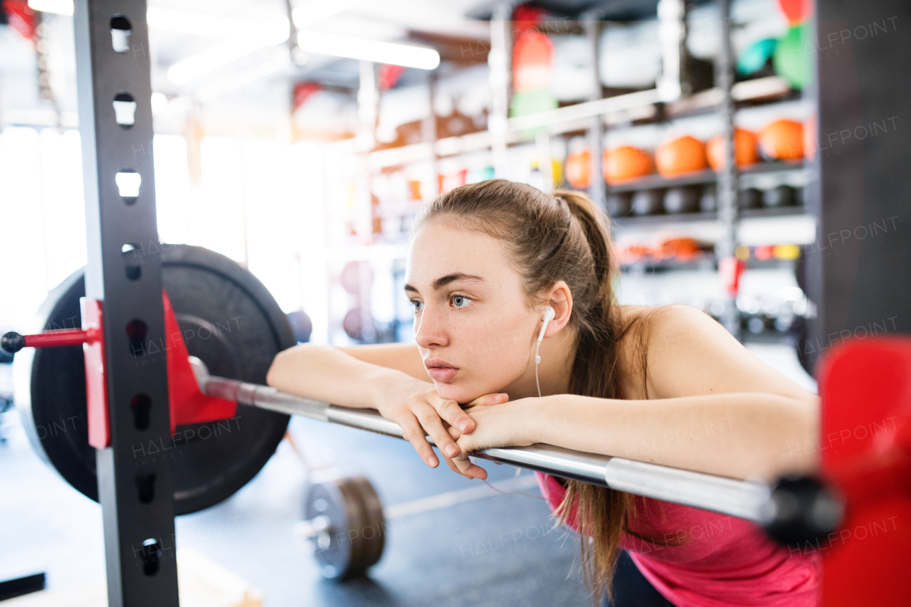 Beautiful young fit woman in gym at the heavy barbell, resting, earphones in her ears, listening music