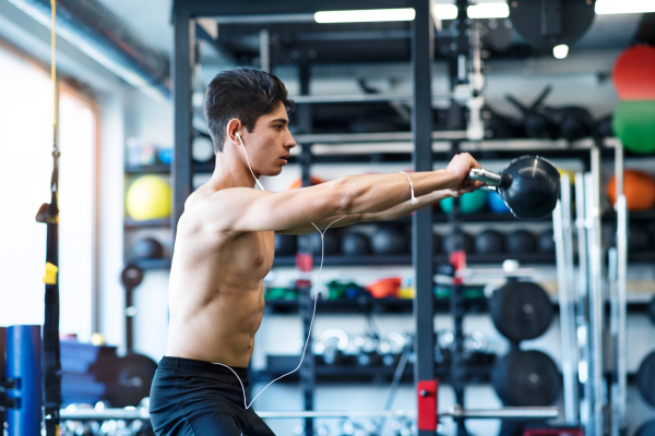 Fit hispanic man doing strength training, doing kettlebell swings in modern gym, earphones in his ears, listening music.