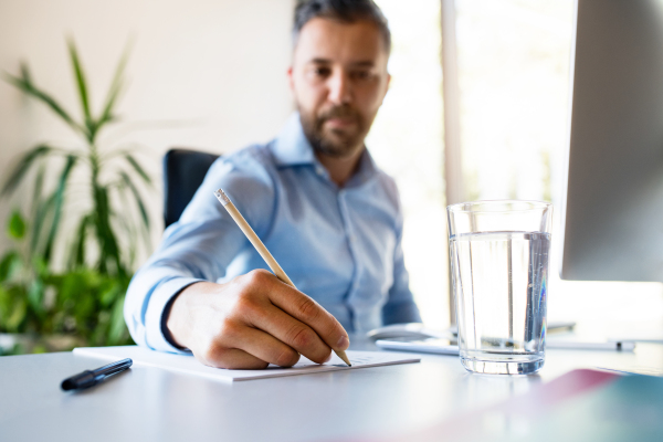 Young businessman in his office, sitting at the desk, computer in front of him, writing something with pencil, glass of water in front of him.