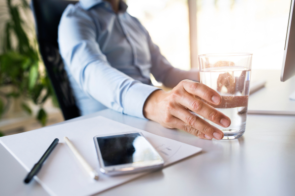 Unrecognizable businessman in his office, sitting at the desk, computer in front of him, holding a glass of water, smart phone next to him.