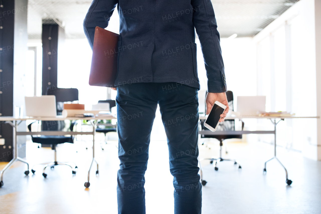 Unrecognizable businessman standing in his office holding smart phone and briefcase. Rear view.