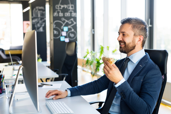 Handsome young businessman in his office, sitting at the desk, computer in front of him, eating his lunch.