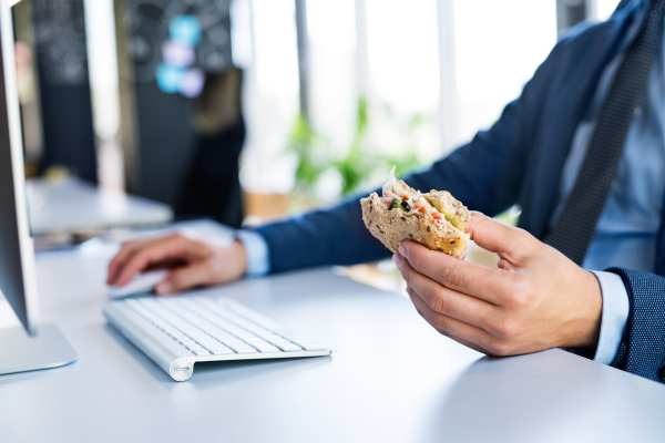 Unrecognizable young businessman in his office, sitting at the desk, computer in front of him, eating his lunch.