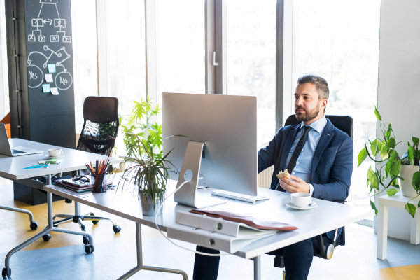 Handsome young businessman in his office, sitting at the desk, computer in front of him, eating his lunch.