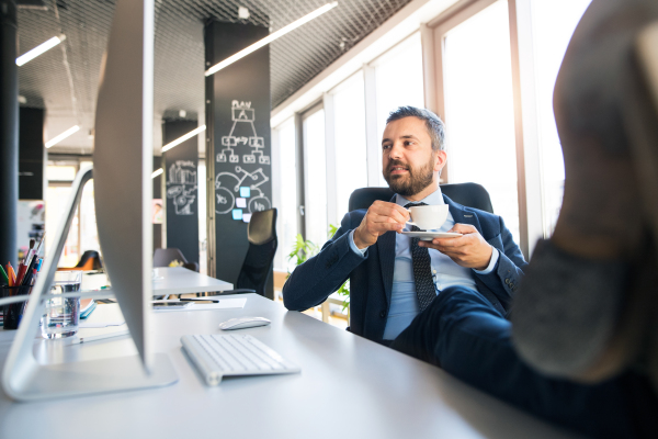 Handsome young businessman working in his office, sitting at the desk, drinking coffee with his feet up.