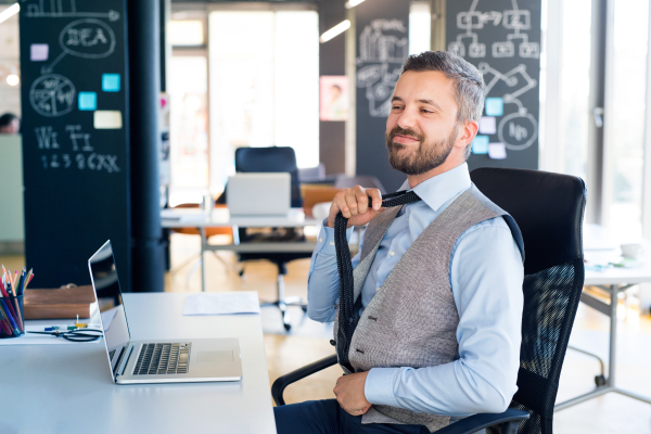 Handsome young businessman in his office, sitting at the desk, laptop in front of him, feeling relaxed.