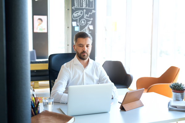 Handsome young businessman working in his office, sitting at the desk, laptop and tablet in front of him.