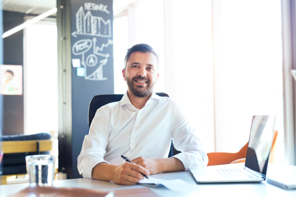 Handsome young businessman in his office, sitting at the desk, smiling.