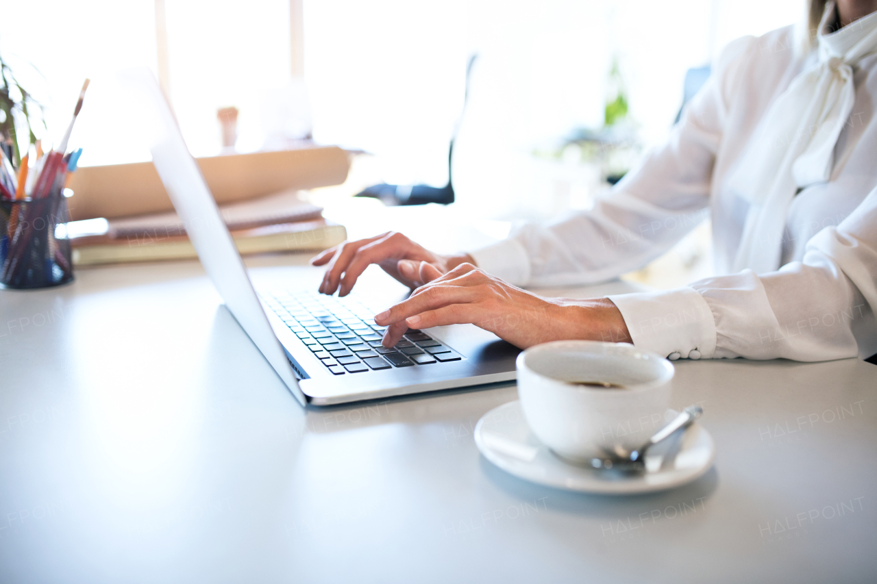 Unrecognizable young businesswoman in her office, sitting at the desk, working on laptop.