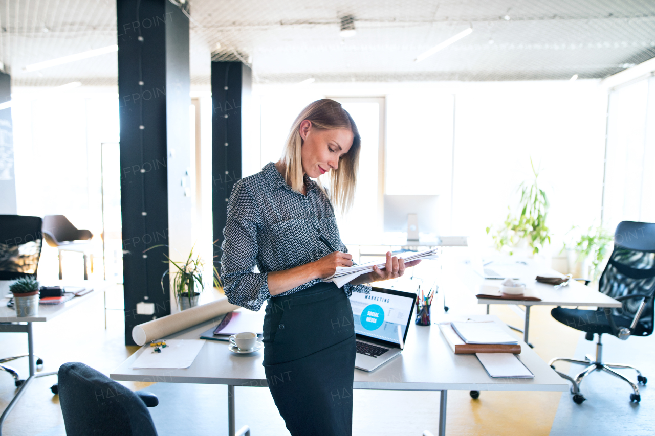 Beautiful young businesswoman in her office at the desk, writing notes or goals into her notebook.